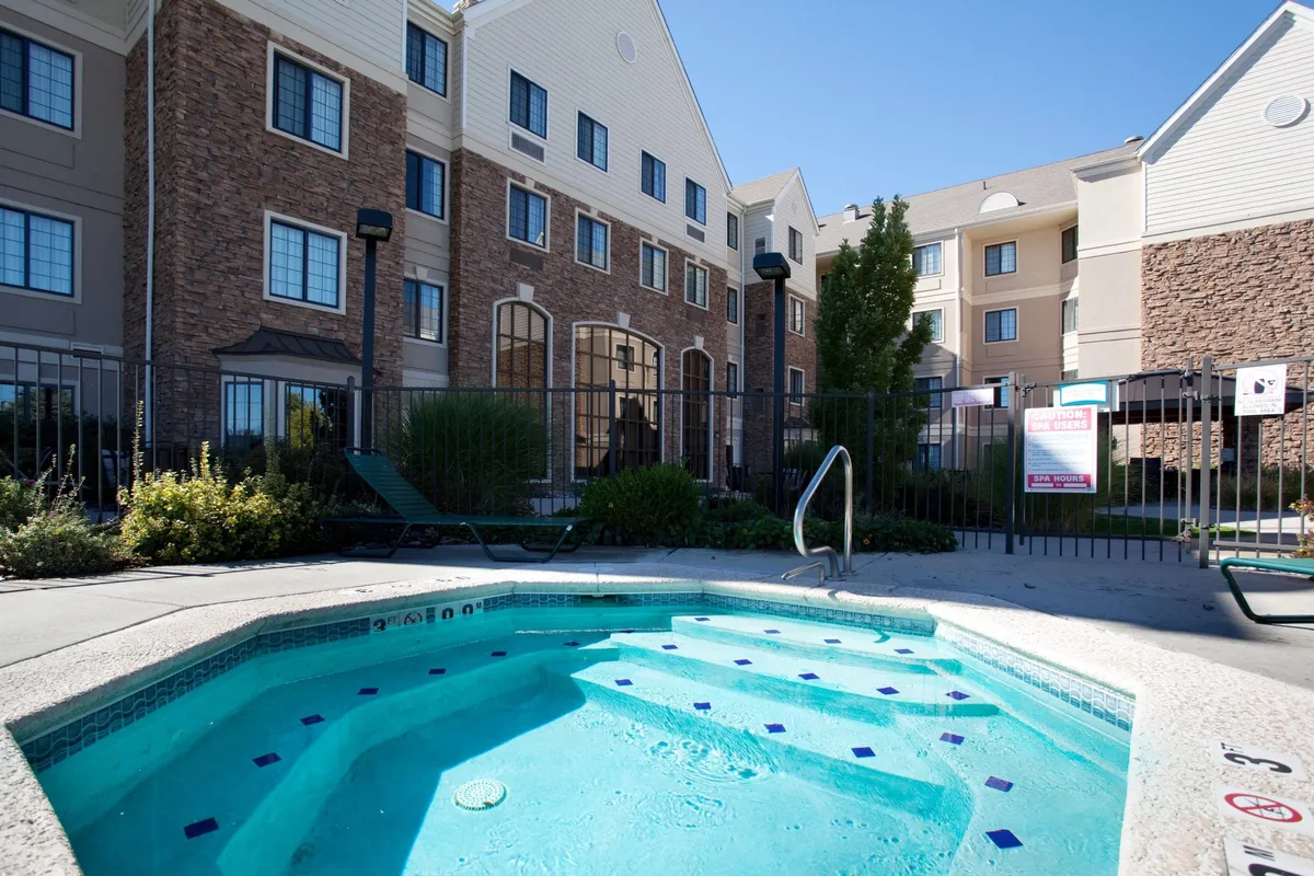 This image captures the inviting outdoor pool area with a hot tub, complemented by attractive landscaping and the building facade in the background. The scene depicts guests relaxing in the hot tub, sunbathing, and enjoying each other's company by the poolside, set in a sunny, family-friendly atmosphere.