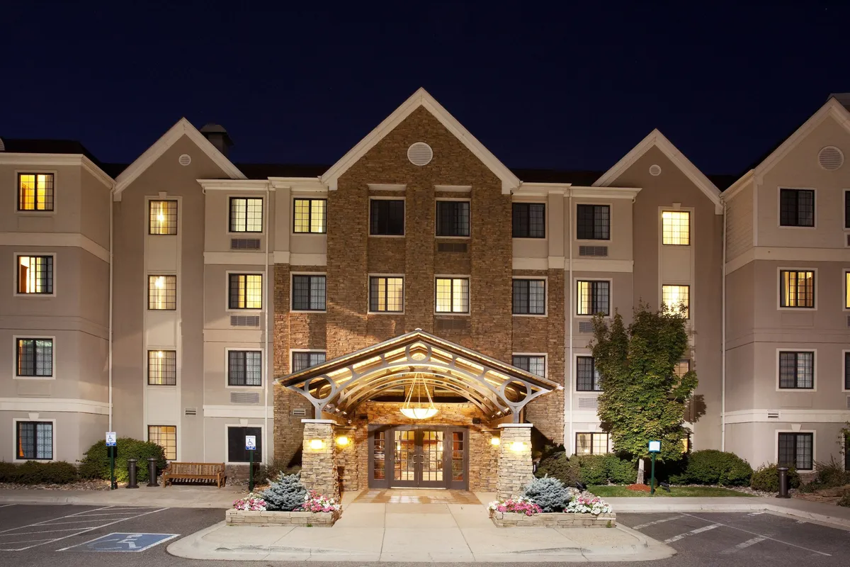 This image shows the exterior of the property with a prominent entrance awning, a stone façade, and warmly illuminated windows. The scene is inviting and suggests both comfort and convenience, with subtle cues of activity like arrivals or gatherings in the lobby just beyond the entrance.
