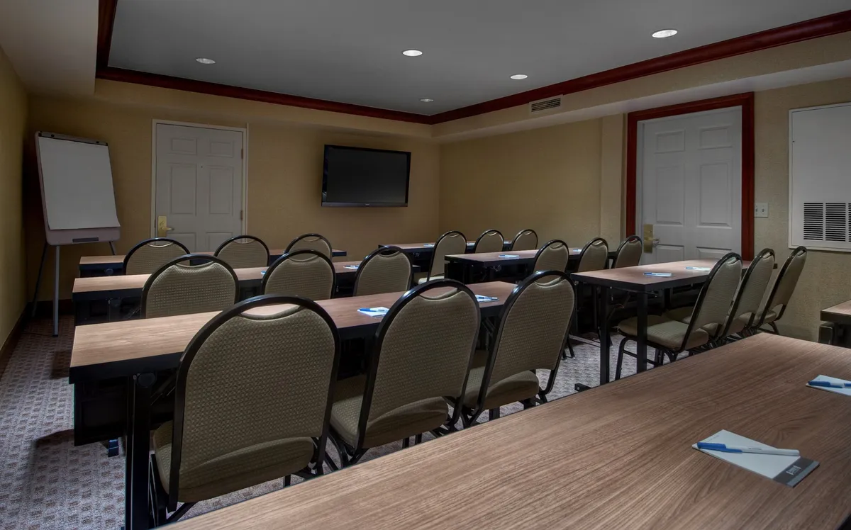 The image depicts an interior meeting space with tables and chairs arranged for a collaborative session. The room features a whiteboard on an easel and a television screen mounted on the wall, suggesting a multifunctional space suited for group gatherings or productive work sessions. The setup feels modern and comfortable, with subtle design elements on the chairs adding a touch of sophistication.