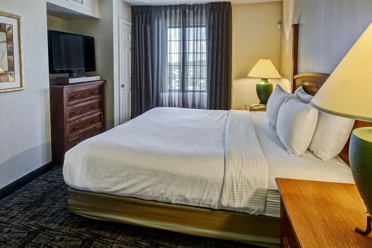 This image reveals a cozy bedroom with a single queen bed, a large window providing natural light, a bedside lamp, and a dresser. The room feels restful and private, ideal for an individual or couple seeking a peaceful night’s sleep.