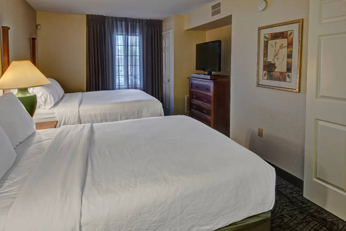 This image presents the primary bedroom, showcasing two neatly made beds with crisp linens, a window draped with curtains that allows natural light to filter in, a wooden dresser, a television, and framed artwork on the wall. The space feels inviting, comfortable, and perfect for families or friends sharing the room.